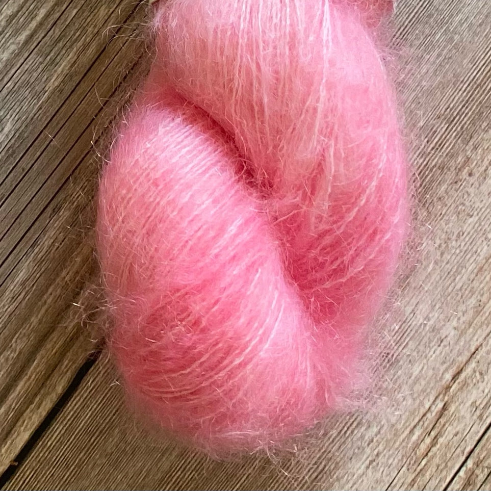 A skein of hand dyed kid mohair and silk yarn in a baby pink color sitting against a wood background