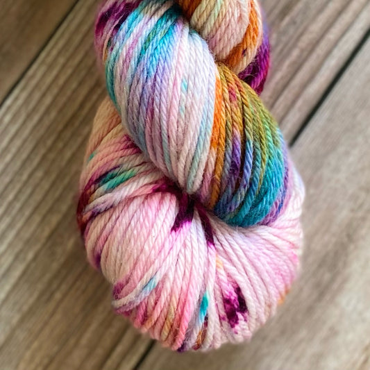 Skein of superwash merino yarn hand dyed in a mix of colors on a soft white background, including turquoise, green, orange, purple and fuschia speckles